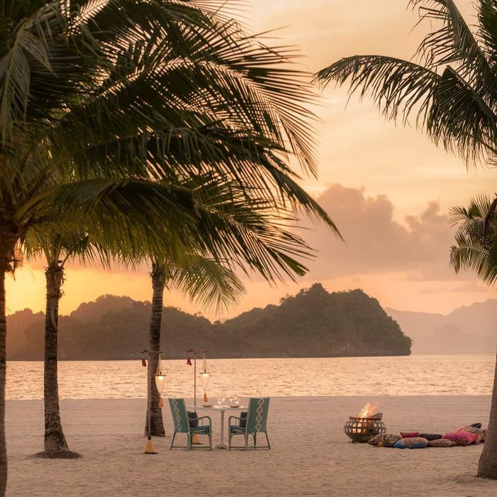 A romantic dinner table set for two on a sandy beach at sunset, flanked by palm trees and a small fire.