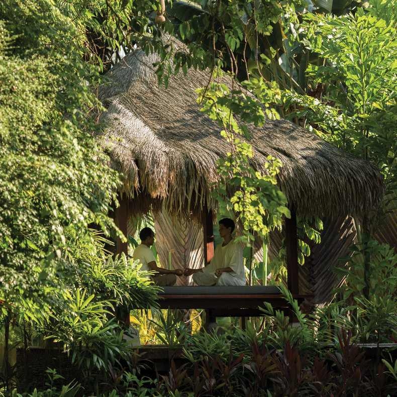 Two people sit in meditation facing each other inside a secluded wooden hut surrounded by lush jungle plants.