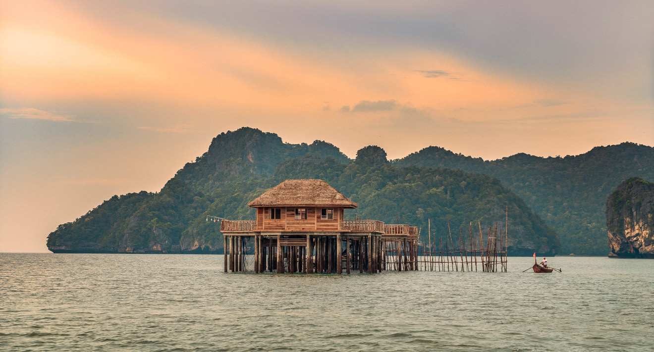 A wooden stilt house sits in the sea with mountains in the background under a warm, hazy sunset sky.
