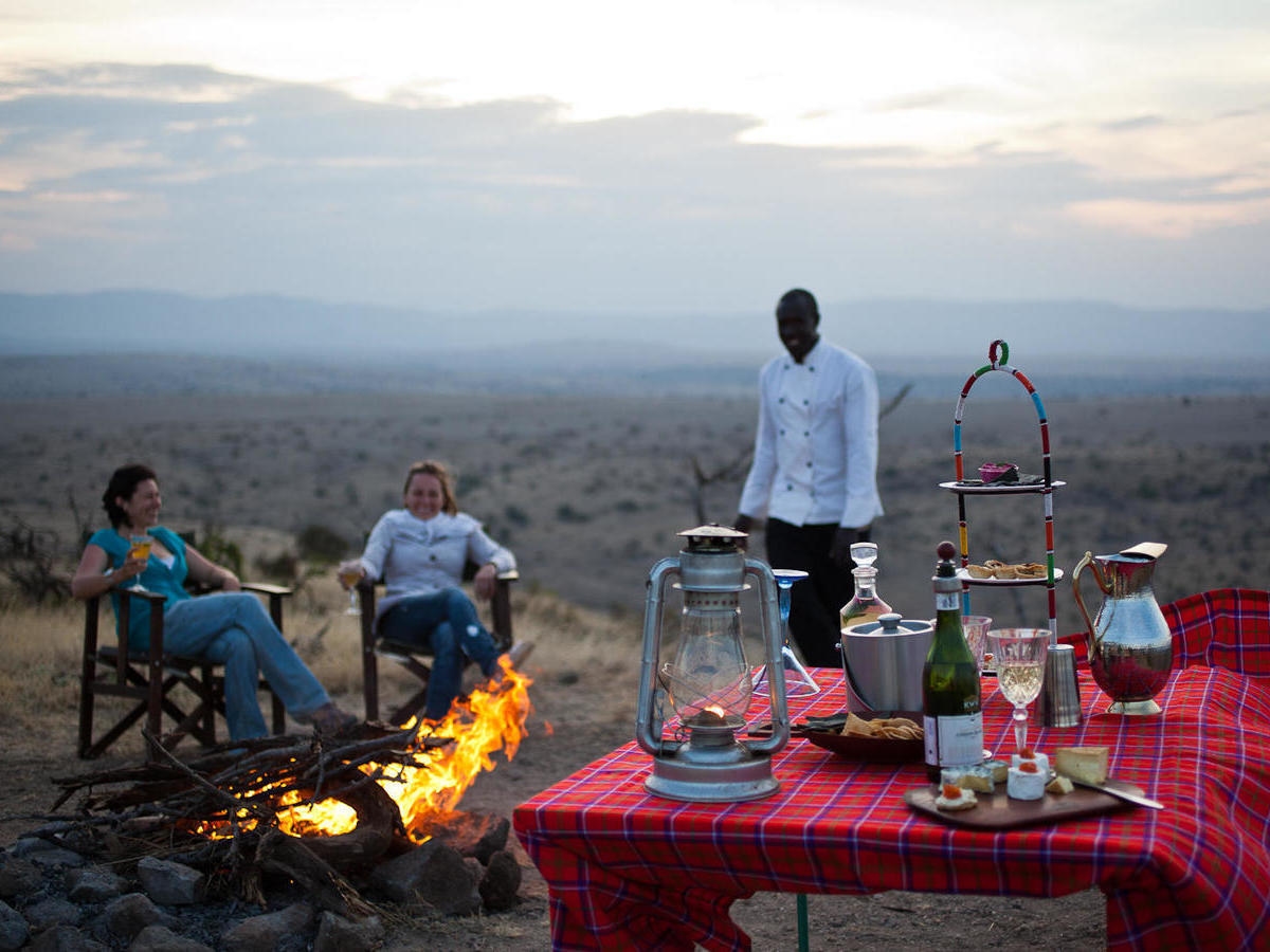 People sitting by a campfire at Lewa House with a table of drinks and snacks overlooking the savanna.