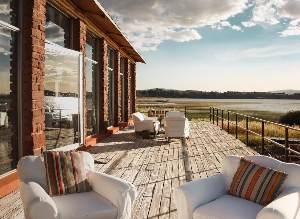 Wooden hotel terrace with white lounge chairs overlooking a calm lake and grassy wetlands under a bright sky.