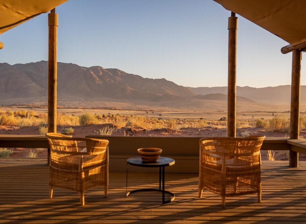 Wicker chairs on a wooden terrace overlooking a desert valley and mountains at sunset.