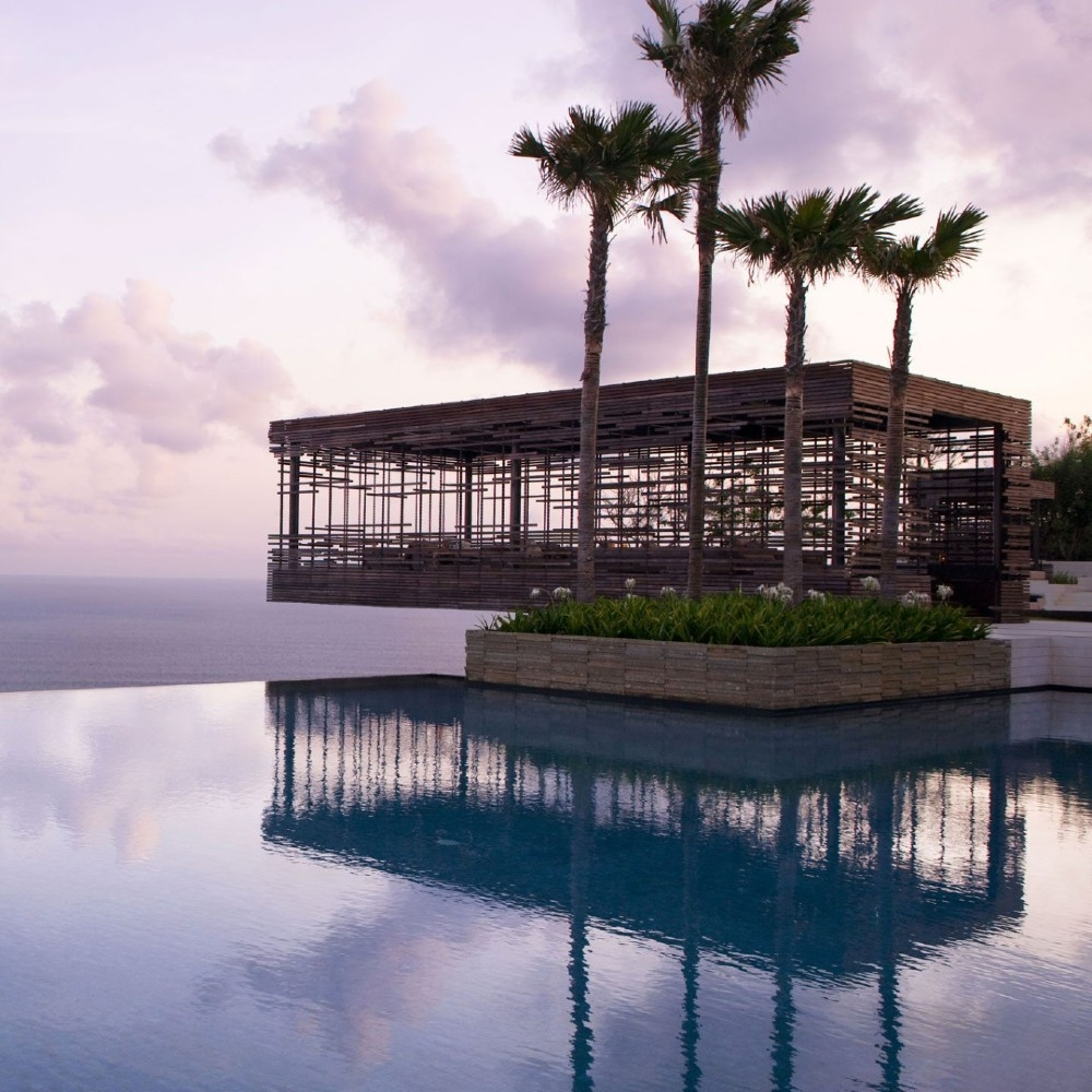 A modern wooden pavilion reflects in a tranquil infinity pool, framed by palm trees and a serene ocean backdrop at sunset.