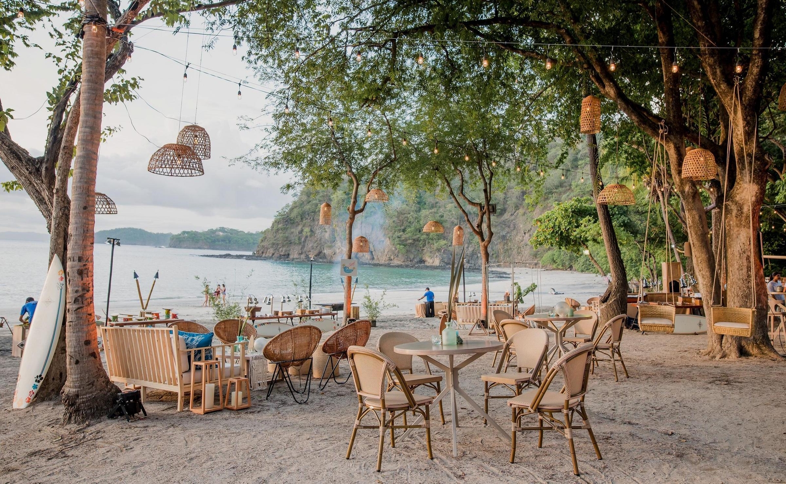 Beachside dining area with hanging woven lamps, wooden furniture, and a surfboard leaning against a tree.