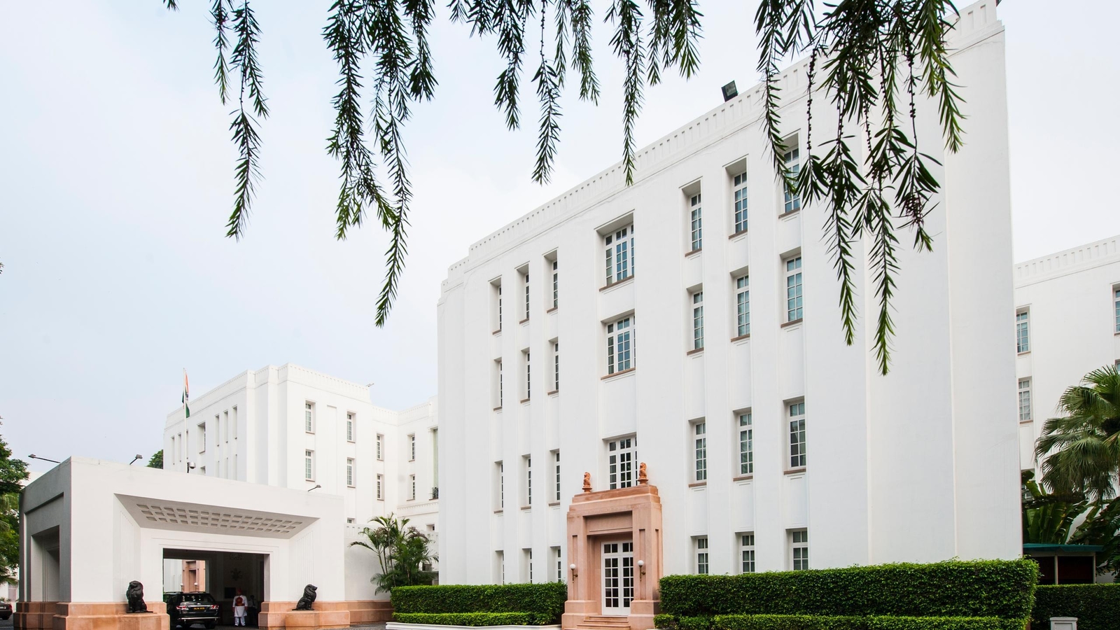 The white exterior of The Imperial Delhi India with a tan stone doorway and manicured green bushes along the base.