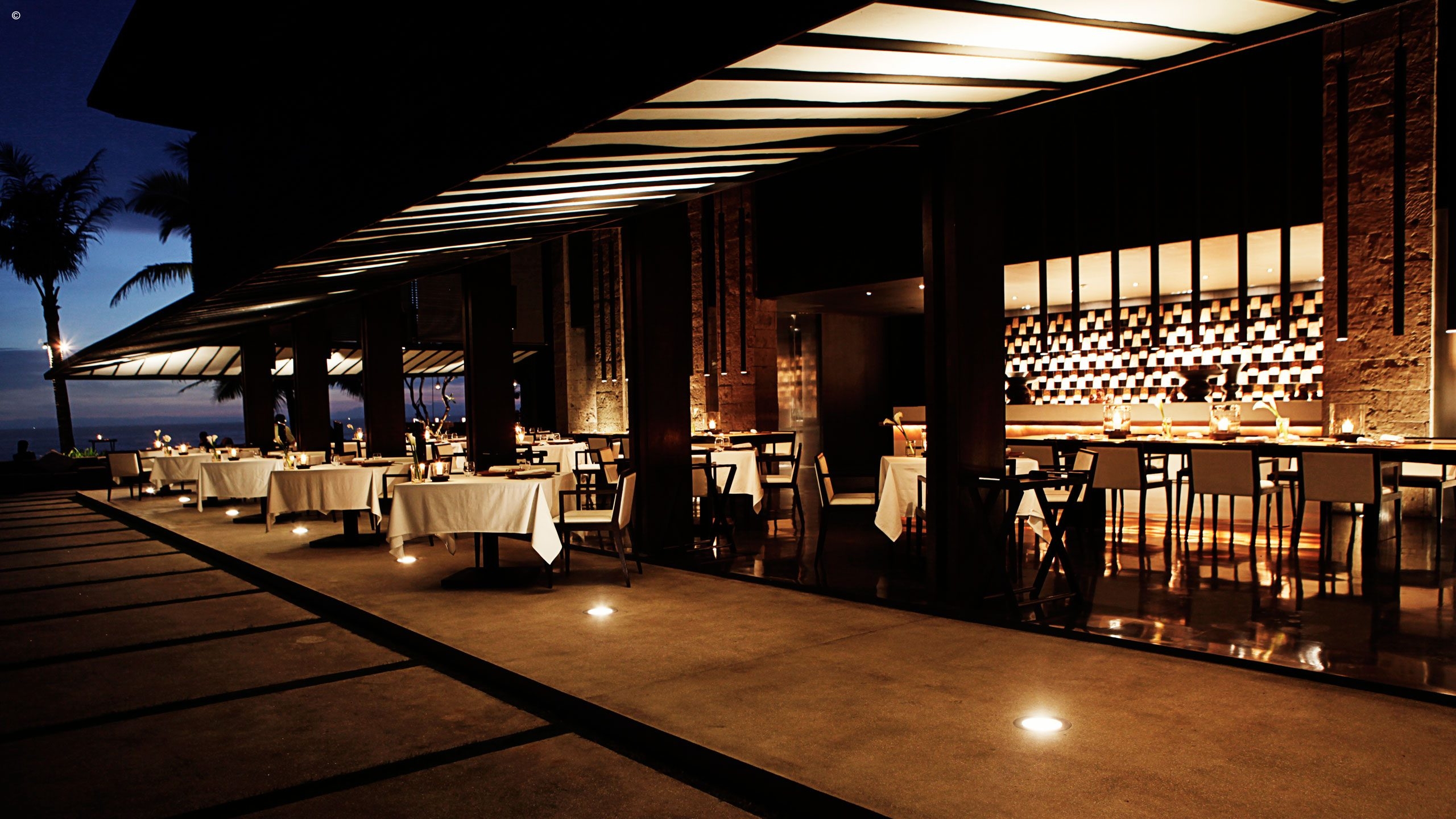 Elegant outdoor dining area at dusk, featuring white tablecloths, soft lighting, and a view of the ocean, framed by palm trees.