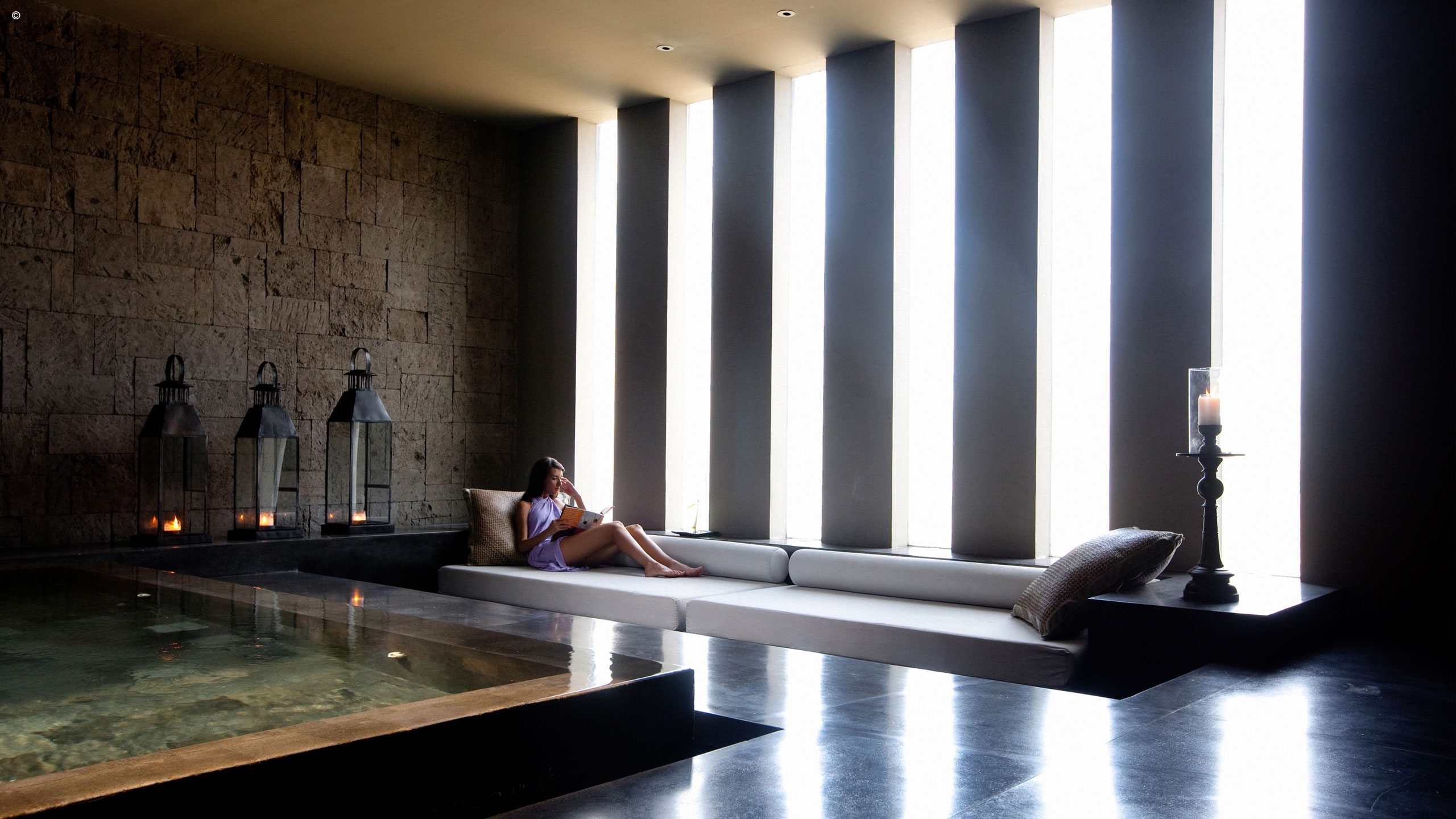 A serene spa setting with a modern design featuring tall windows, candles, and a cozy seating area next to shimmering water.