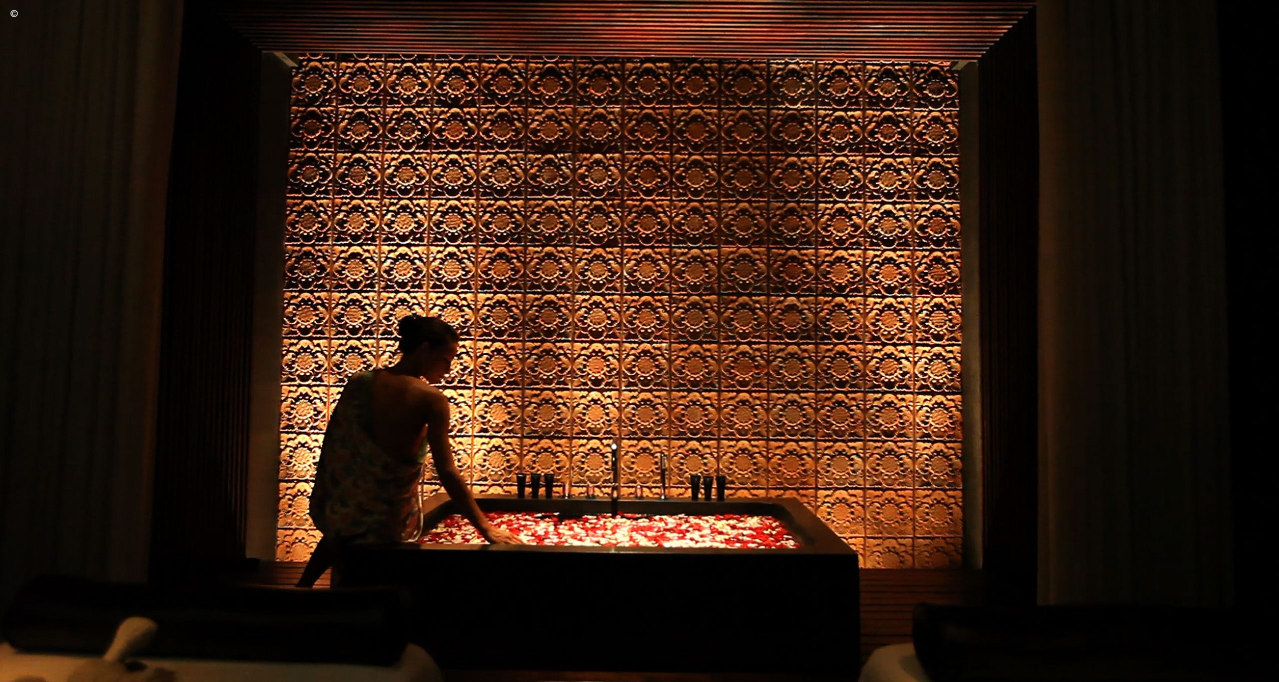 A person leans over a luxurious bathtub filled with rose petals, against a beautifully illuminated patterned wall.