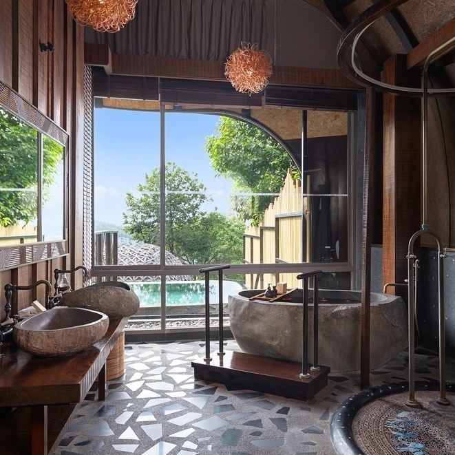 The bathroom of one of Keemala's villas featuring a stone bathtub, two sinks and a shower area overlooking a pool.