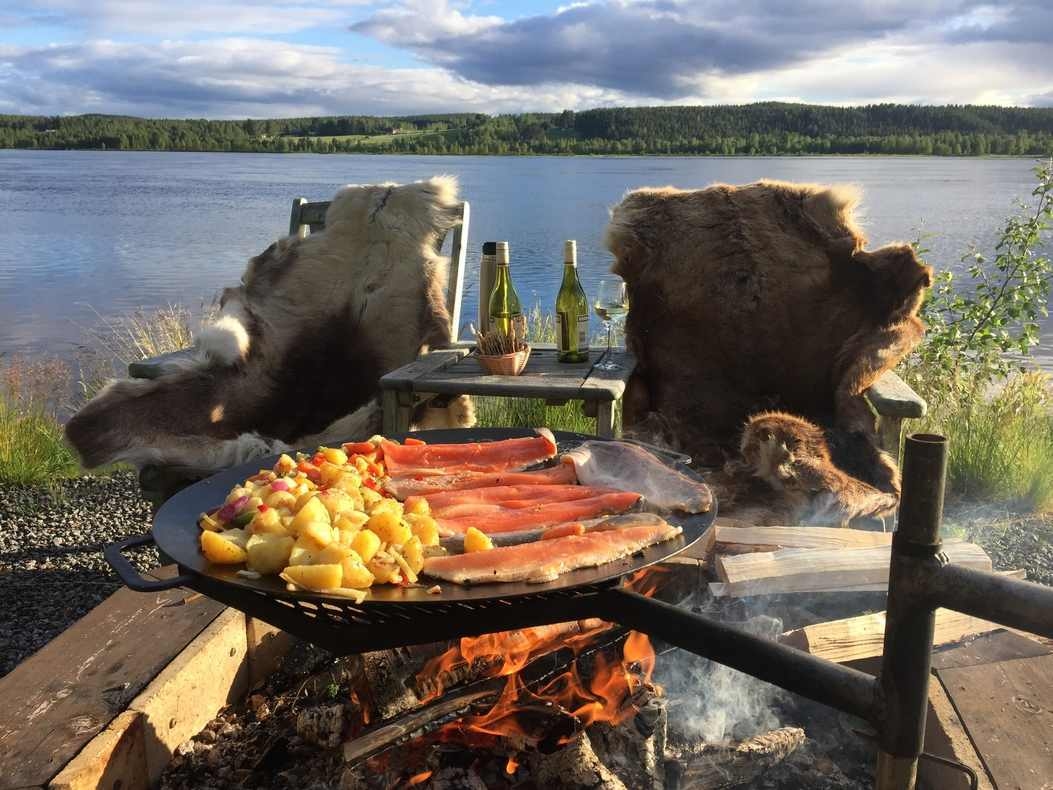 Salmon fillets and potatoes grilling on a fire pit by a river with wine on a table nearby.