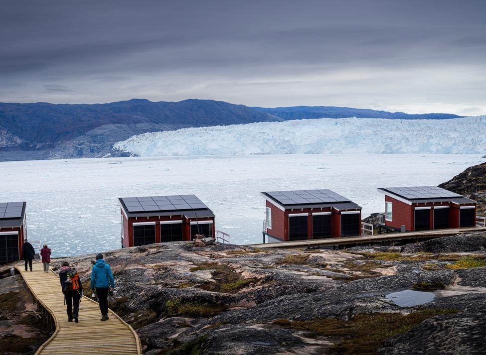 Four red wooden lodges at Eqi Glacier Lodge on a rocky cliff overlooking a glacier in Greenland