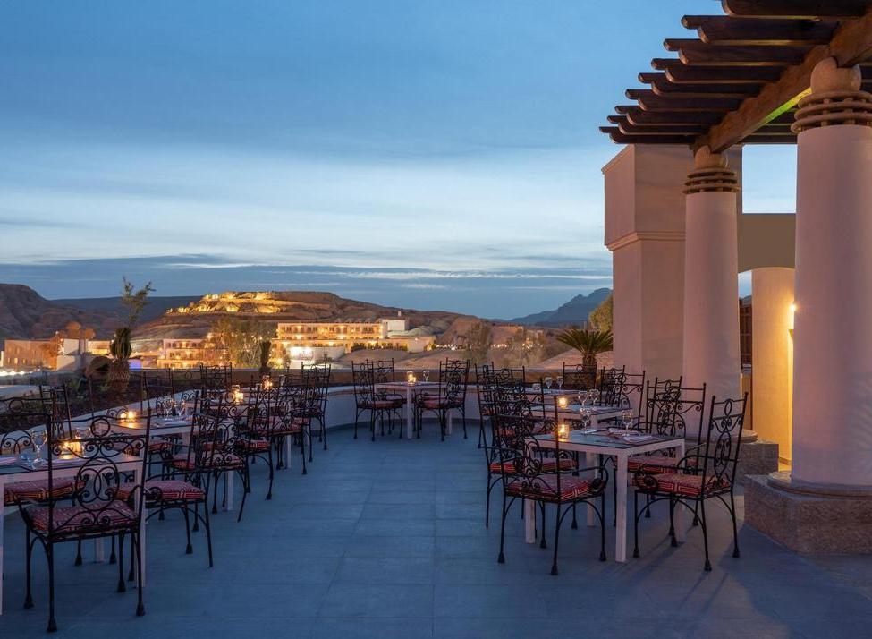 An outdoor restaurant terrace with set tables and candles overlooking a lit-up hillside town at dusk.