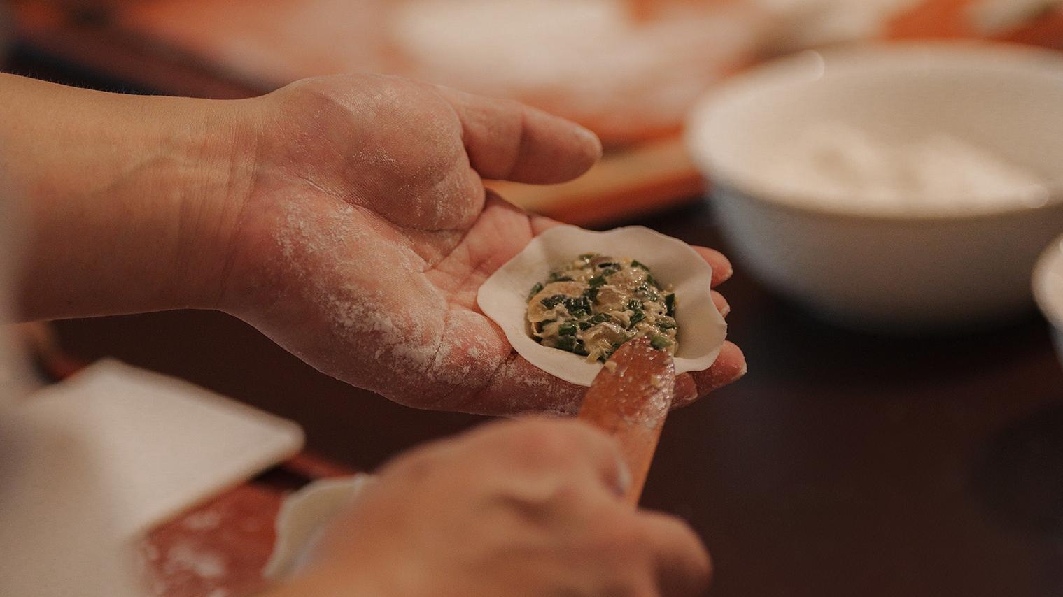 Hands preparing a dumpling with filling, dusted with flour over a wooden table.