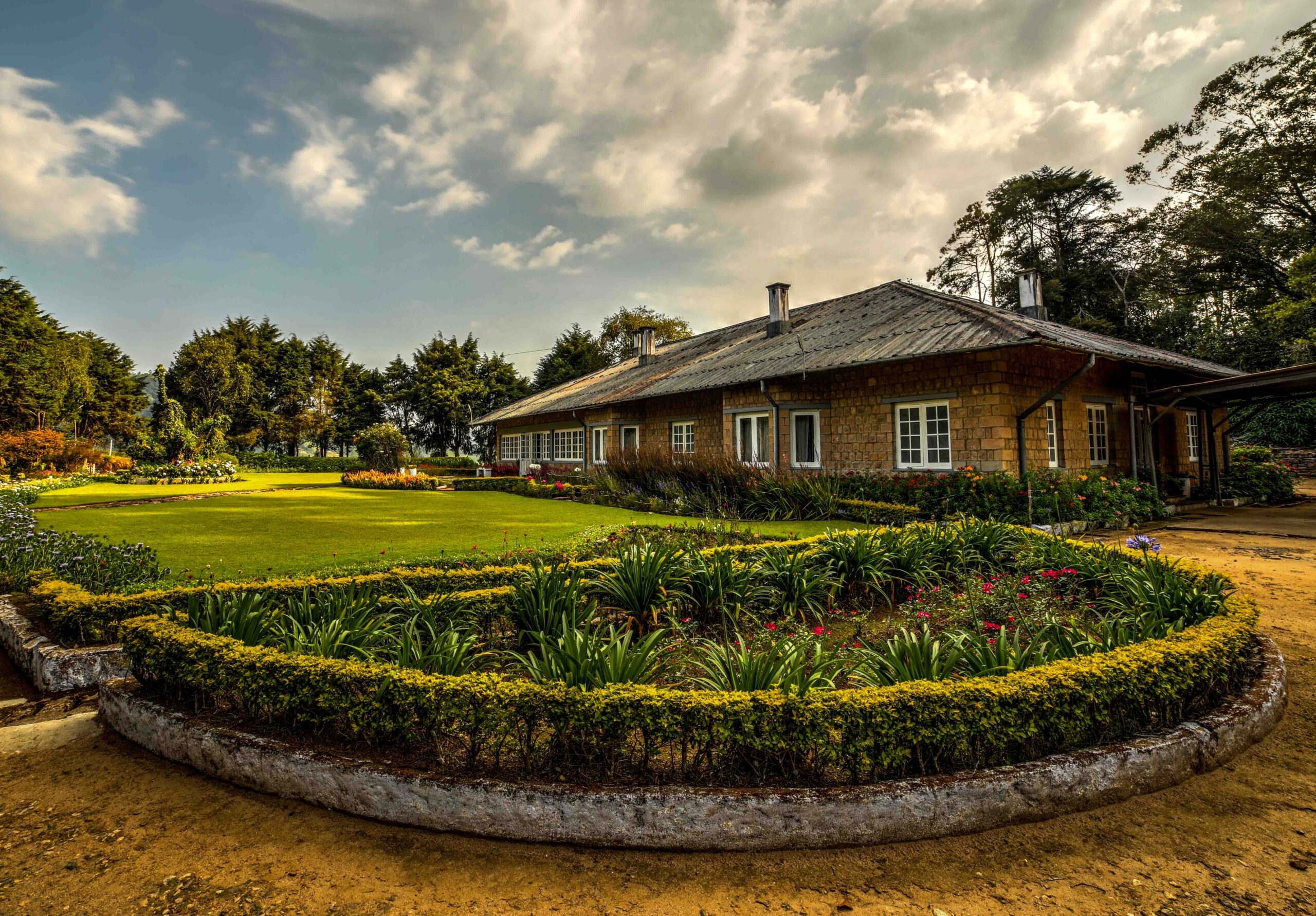 Rustic stone cottage with a lush garden and manicured lawns under a cloudy sky.