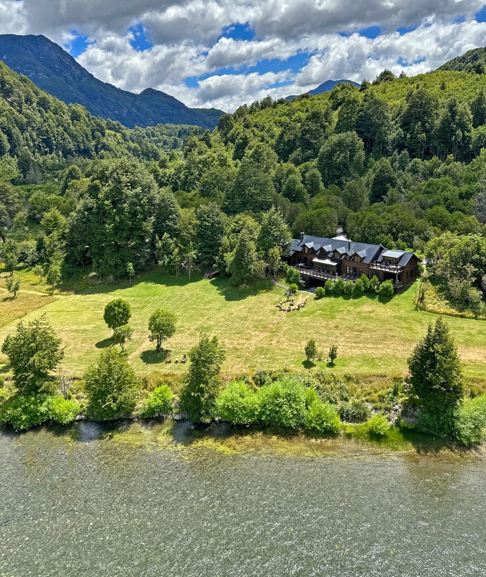 Aerial view of a large wooden house beside a river, with surrounding greenery and mountains in the background.