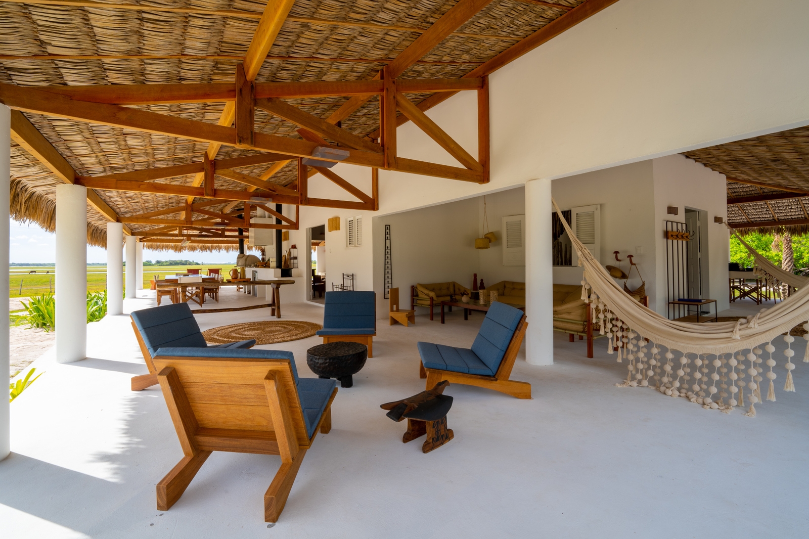 Four blue lounge chairs on an open-air deck with wooden beams, a hammock, white walls and a thatched roof, and a view to grass outside.