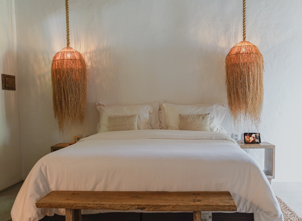 Interior shot of a double bed with white sheets and beige pillows framed by two hanging lamps with dried grass for lampshades, and a wooden bench in front of the bed.