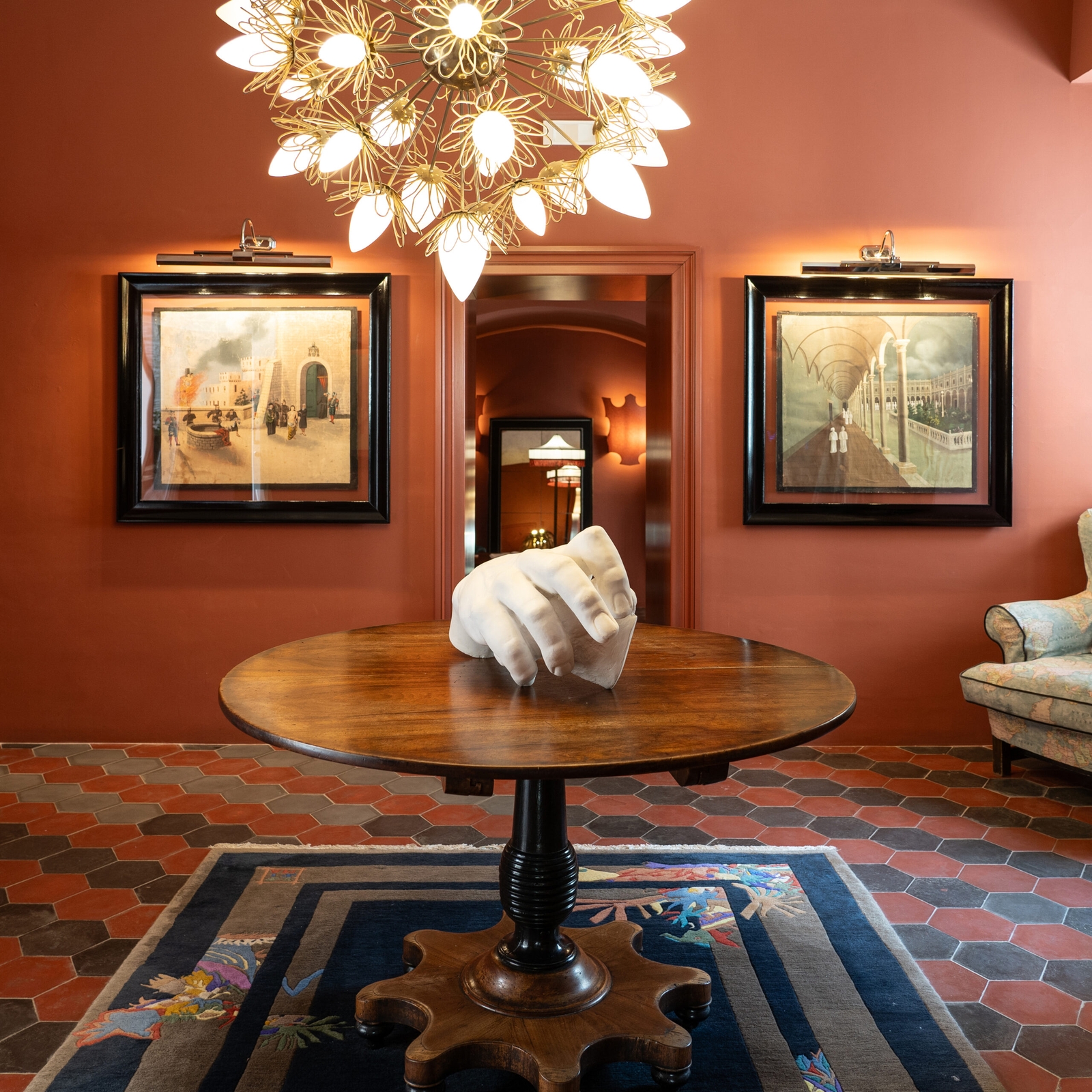 Interior entryway with red walls, a chandelier with bare bulbs hanging over a table with a sculpture of a hand, and two paintings on the back wall.