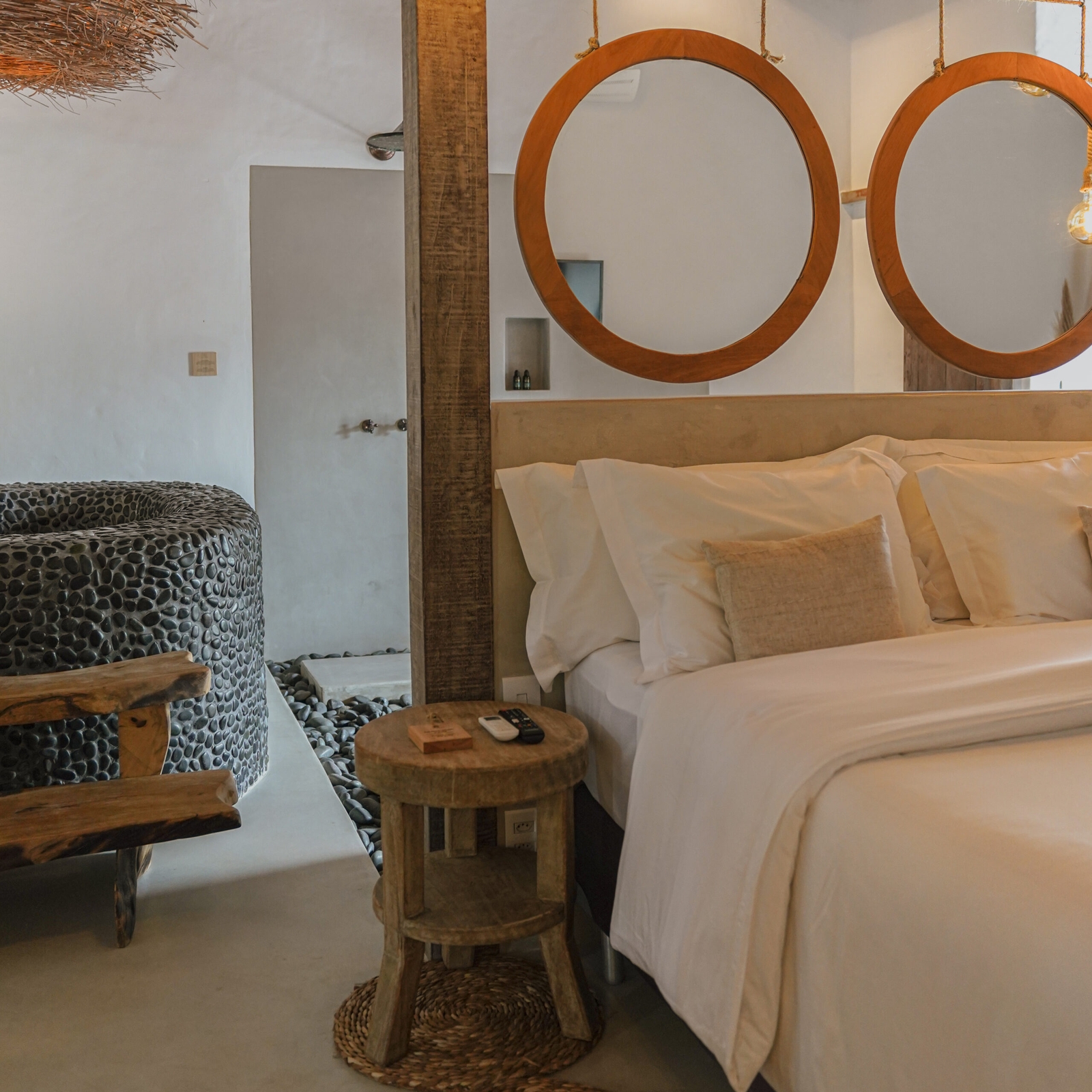 Interior of a bedroom - a double bed with white and beige sheets, a small wooden bedside table, grey-wood beams, and in the background a round soaking tub made of concrete and stones.