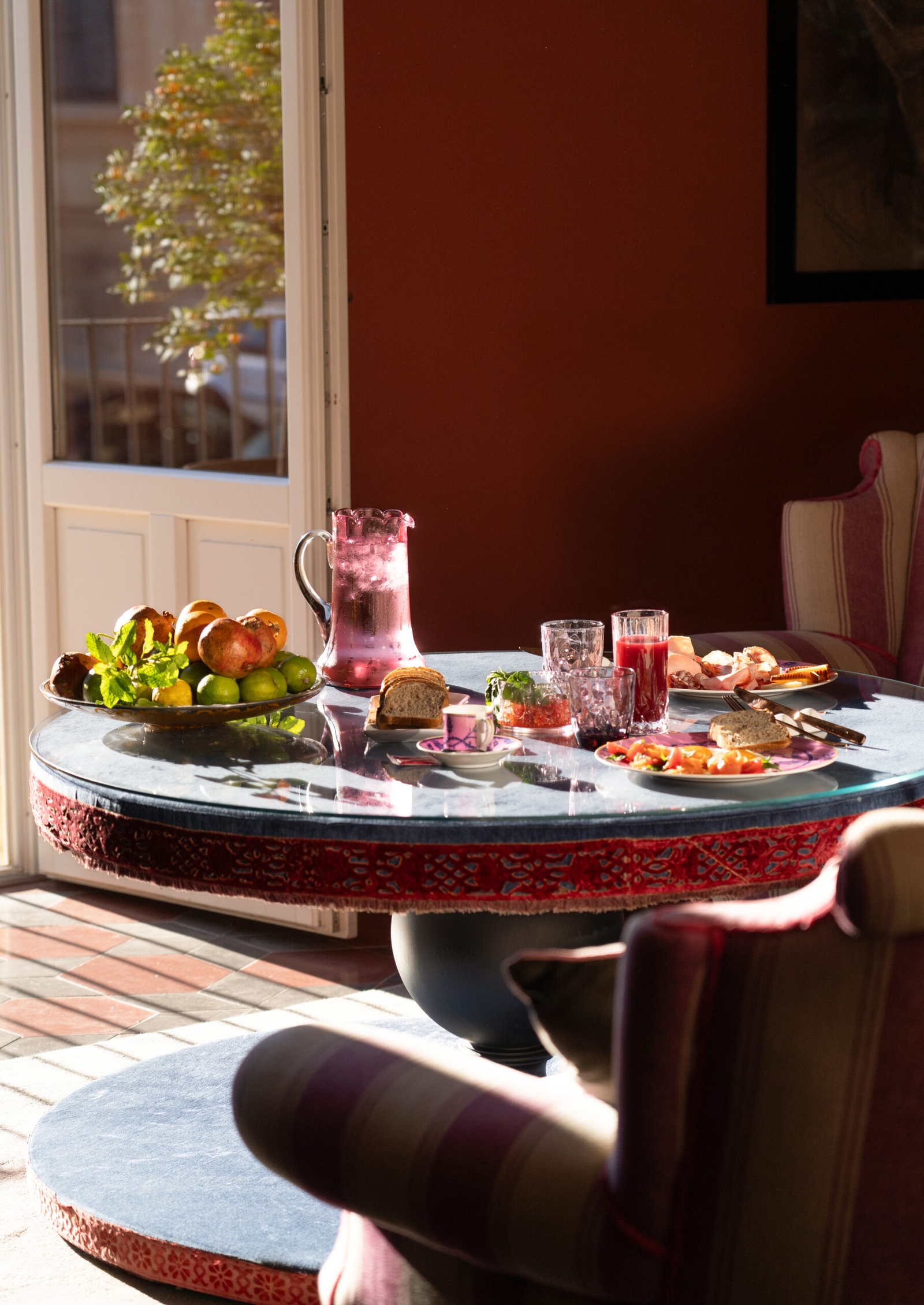 Interior shot of a breakfast table with a jug of juice and plates of fruit and pastries. In the background, a door opens onto a terrace.