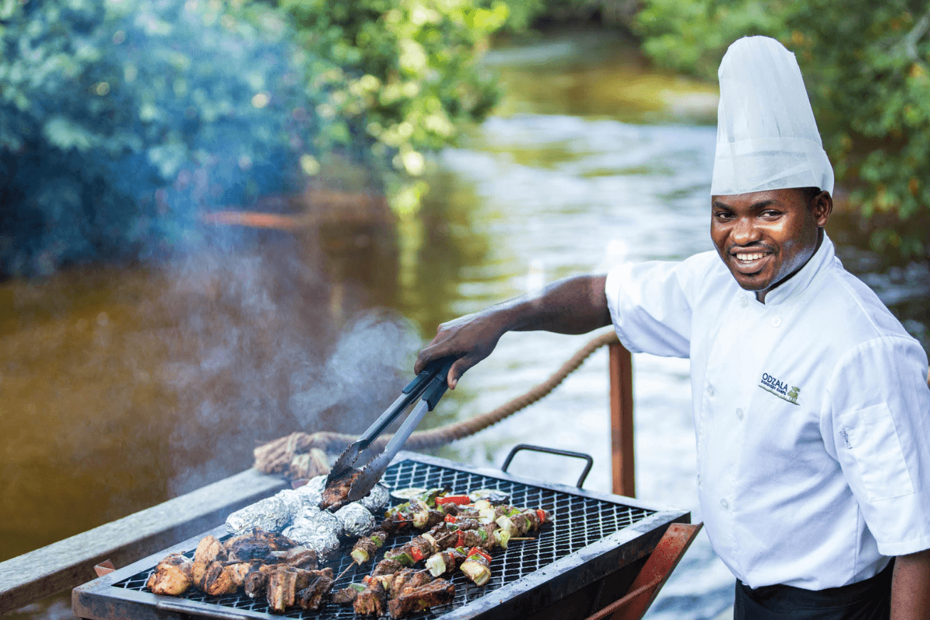 A smiling chef tending to the food on a smoking grill beside a river
