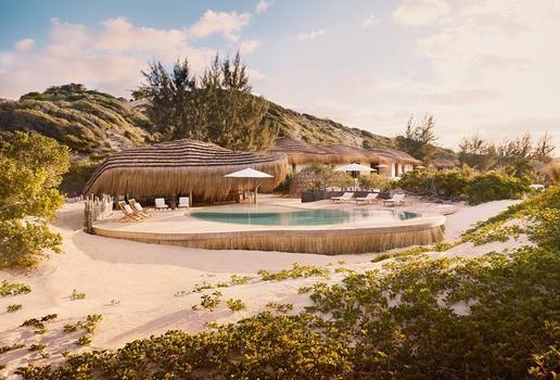 A curvy swimming pool in front of thatched-roof buildings on a sandy beach landscape under a soft sky.