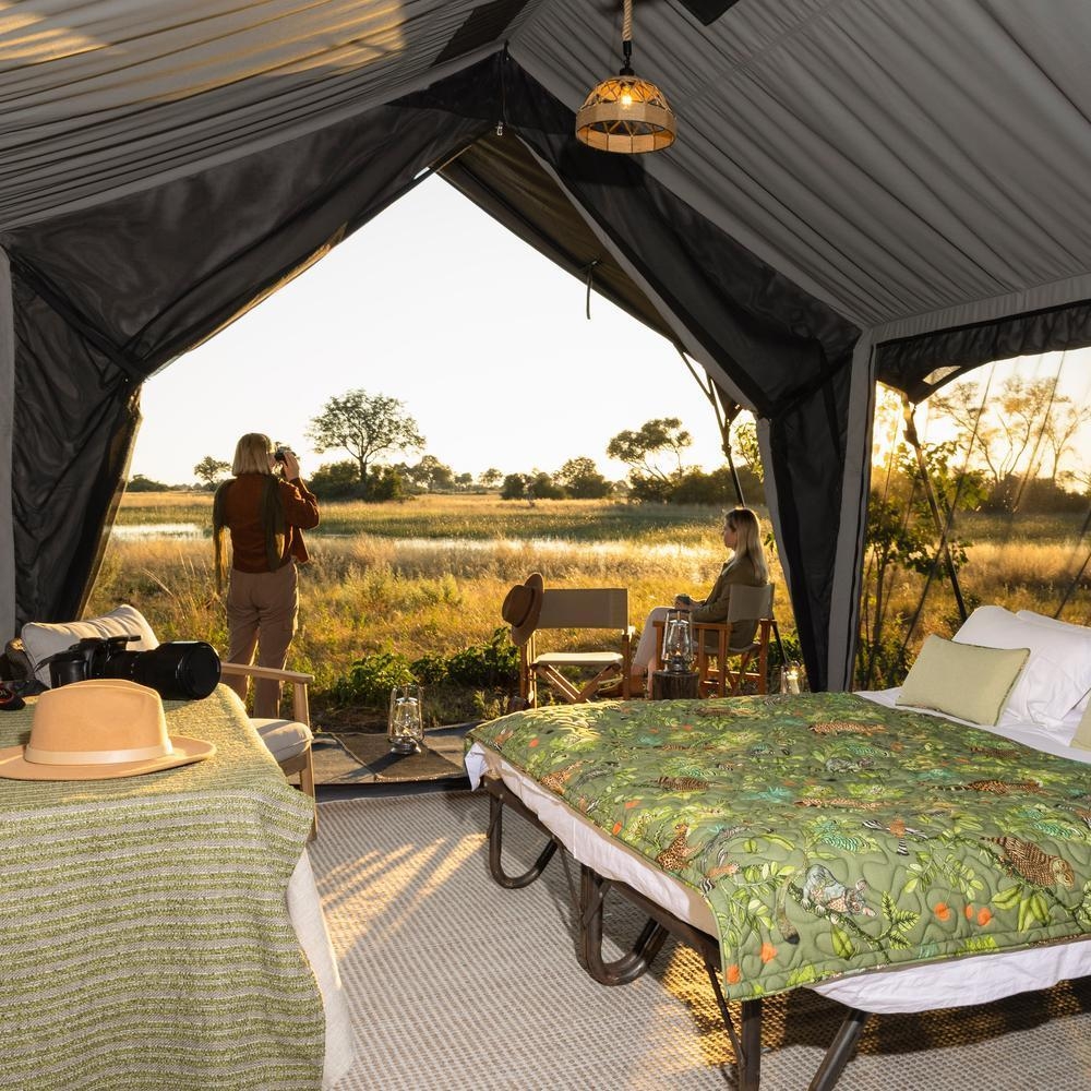 Inside a safari tent showing a bed with an animal print duvet and a person looking at the landscape with binoculars.