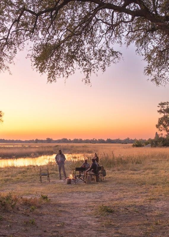 A group of people sitting in camp chairs around a small fire next to a river at sunset.