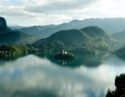 Aerial view of Lake Bled in the Alps, with a small church on an island in the middle of clear still lake surrounded by mountains and thick forests