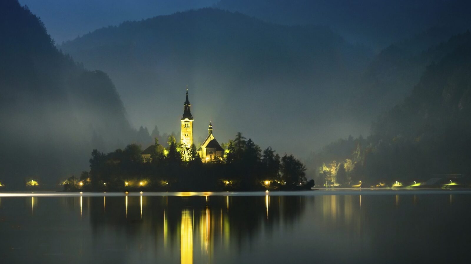 Nighttime view of small island with church on it in Lake Bled, Slovenia. The church is illuminated by yellow light