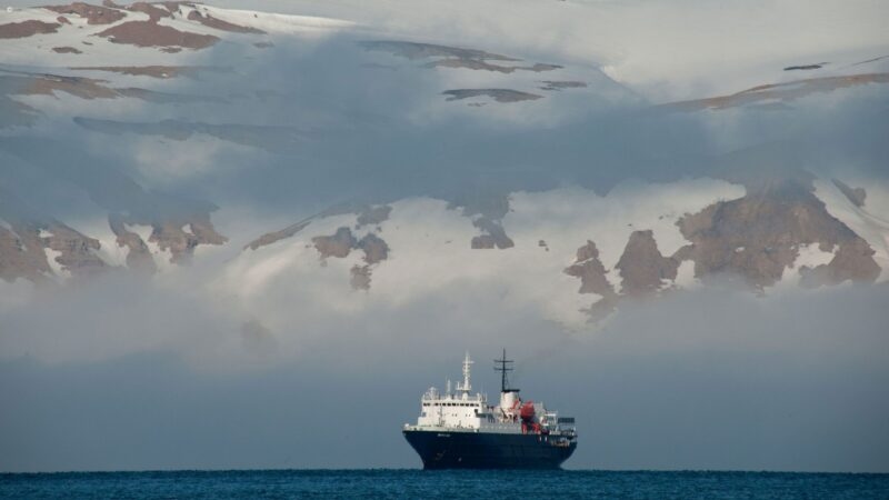 A white and black ship on the ocean with large, snowy, cloud-covered mountains in the distant background.