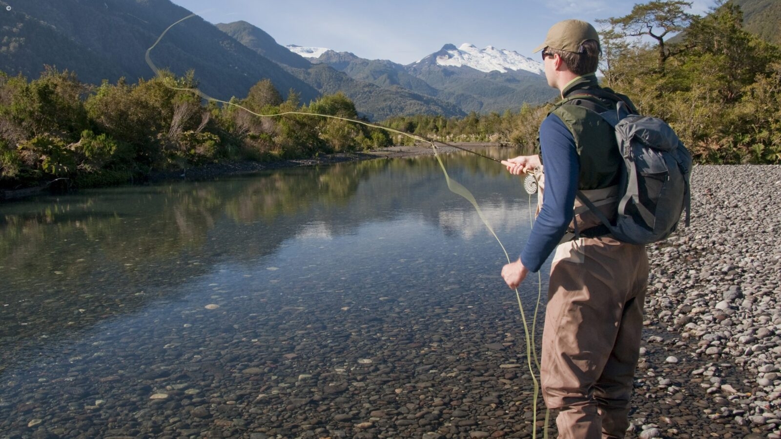A man in brown trousers and a navy blue shirt with a green hat and backpack casting a long fishing line in a clear river with snowcapped mountains in the background