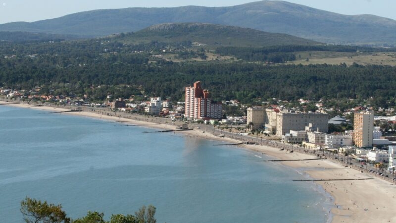 High-angle panorama of a city coastline and sandy beach with a large mountain under a clear blue sky.