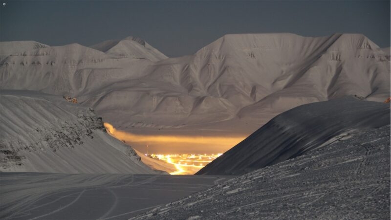 Snow-covered mountain peaks at night with a distant town glowing brightly in a dark valley between slopes.