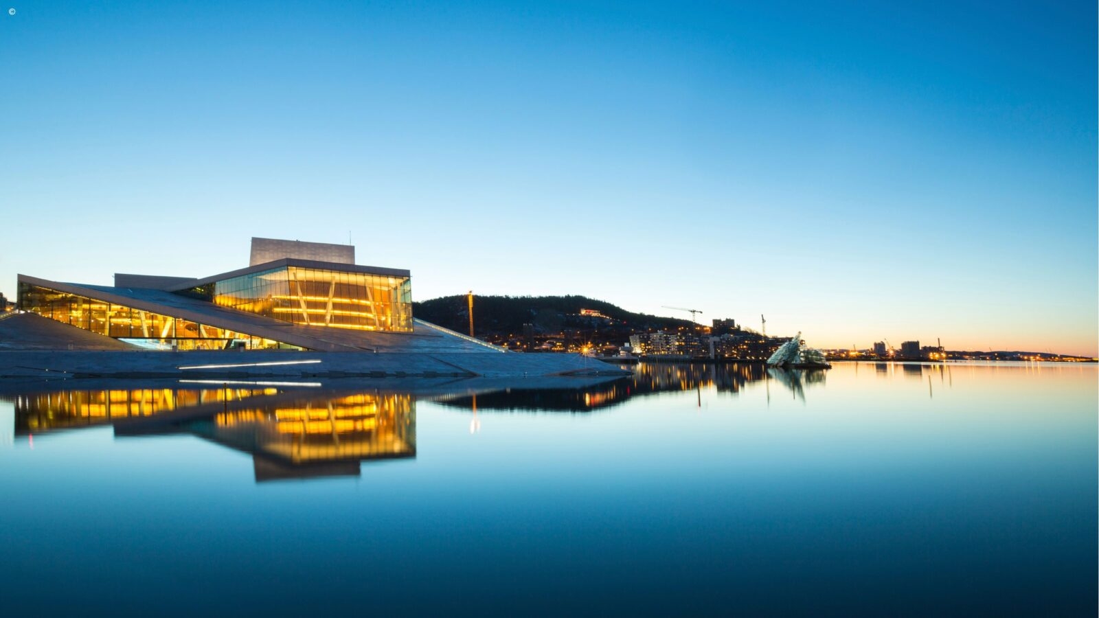A modern architectural building with glowing interior lights reflecting on still water under a clear dusk sky.