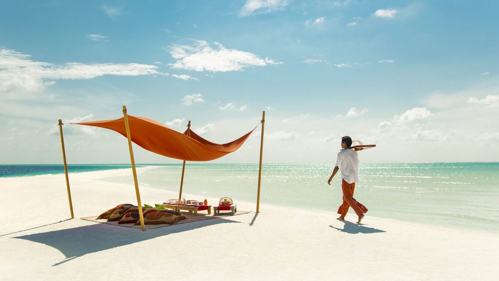 A mean carrying a tray towards a picnic setup under a shelter on a white sandy beach by the sea