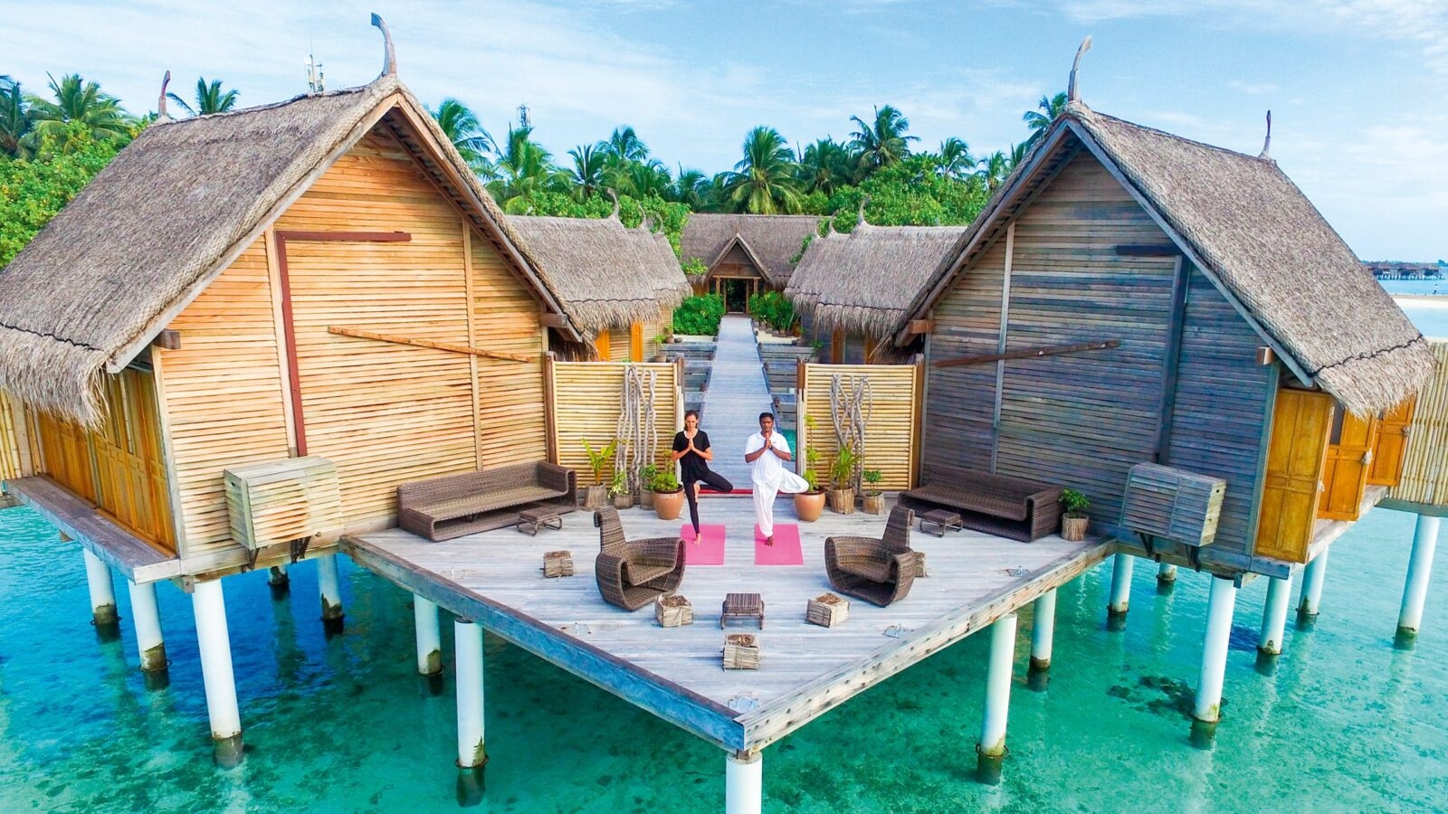 Two people practising yoga on pink mats on a wooden stilted jetty next to overwater bungalows on a reef