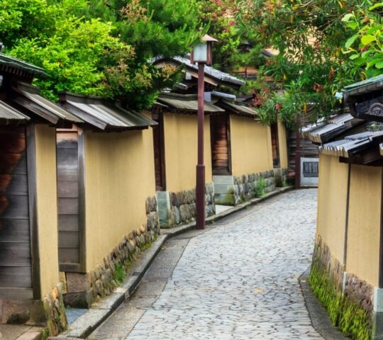 A narrow stone path winding between traditional Japanese buildings with yellow walls and dark tiled roofs.