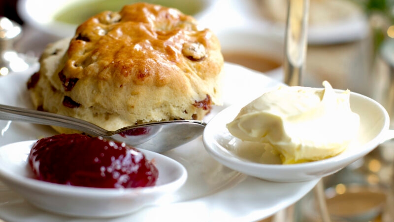 A golden-brown fruit scone served with a side of red strawberry jam and thick clotted cream on white chinaware.