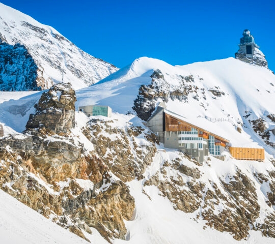 Modern wooden building built into a snowy, rocky mountain slope under a bright blue sky.