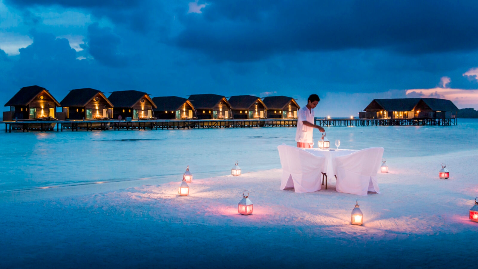 A romantic candlelit doinner set up on a white sand beach with overwater bungalows in the background