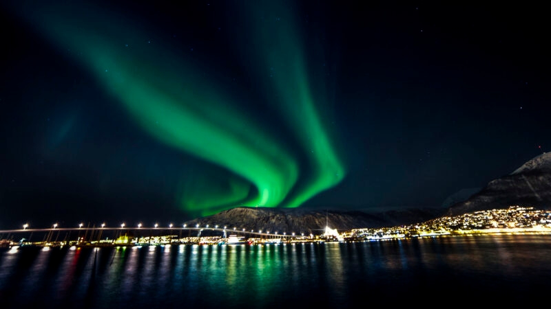 Bright green northern lights waving in the dark sky over a glowing city waterfront and long bridge.