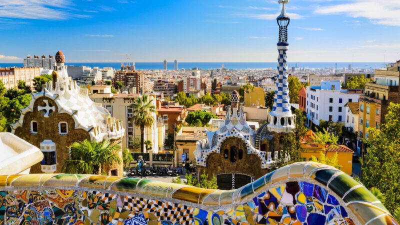 View of Park Guell's mosaic walls and iconic buildings with the Barcelona city skyline in the background.