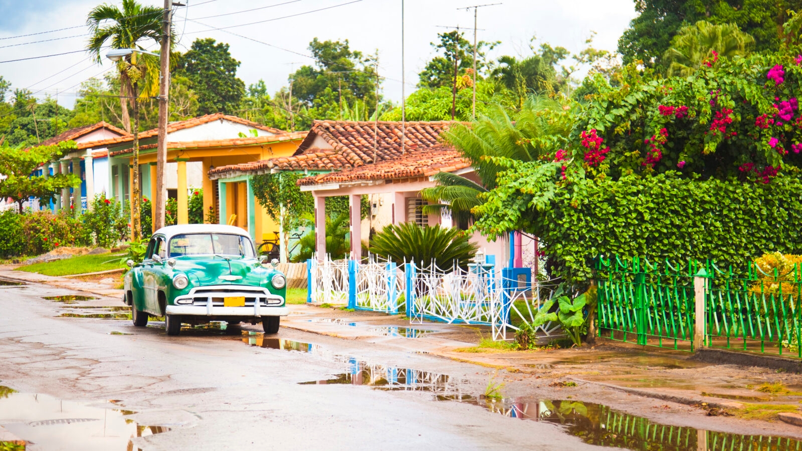 A vintage green and white car parked on a residential street with puddles and colorful houses.