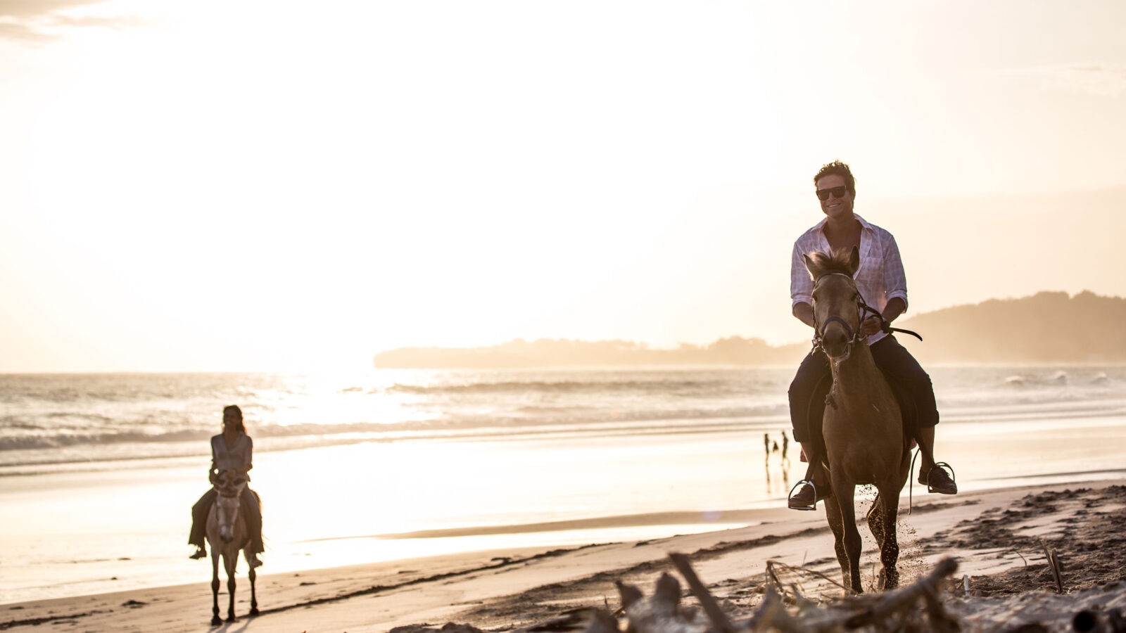 A man and woman ride horses along a sandy beach during a bright sunset with ocean waves in the background.