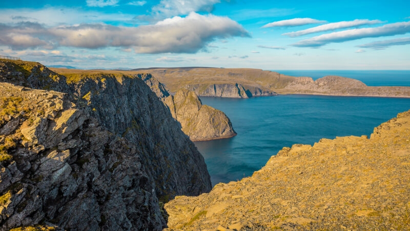 Wide view of steep, rugged coastal cliffs meeting the blue ocean under a sunny sky with soft clouds.