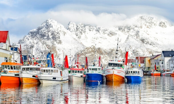 Colorful fishing boats docked in a harbor in front of massive snow-covered mountains under a cloudy sky.