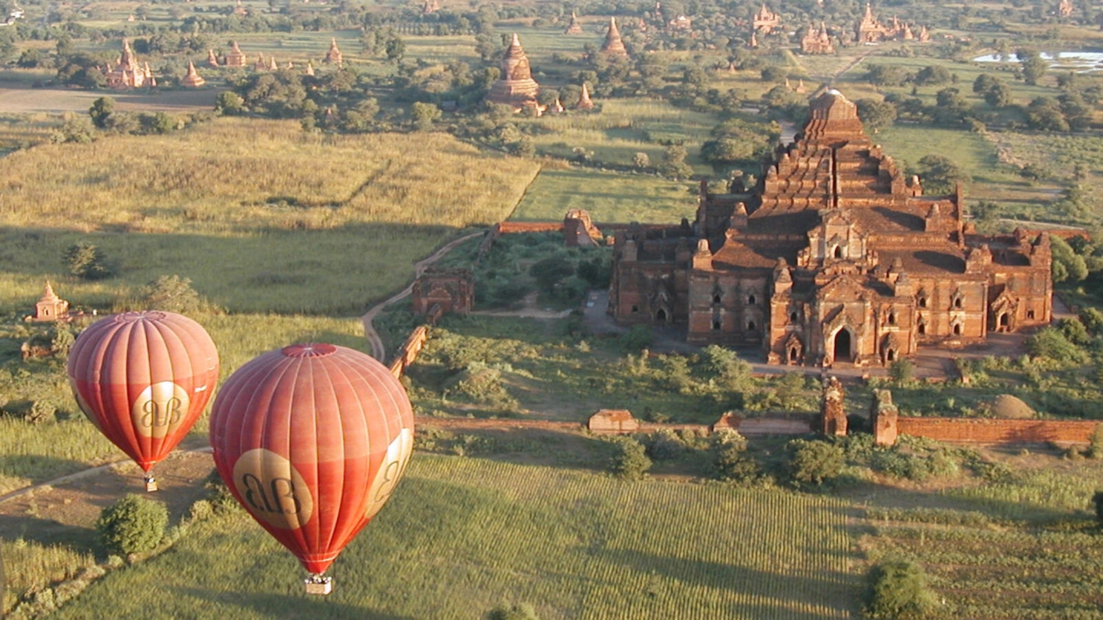High-angle view of the Dhammayangyi Temple in Bagan with two red hot air balloons flying over green fields.