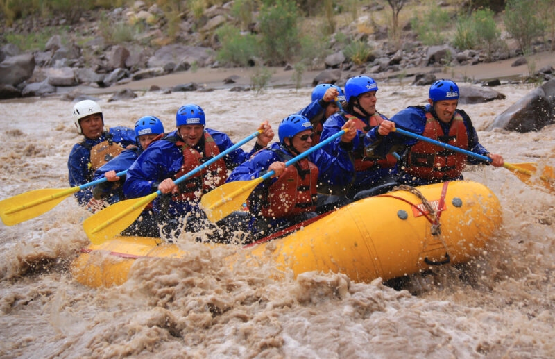 A group of eight people white-water rafting on a turbulent brown river, paddling a yellow inflatable boat. Luxury Argentina holidays.