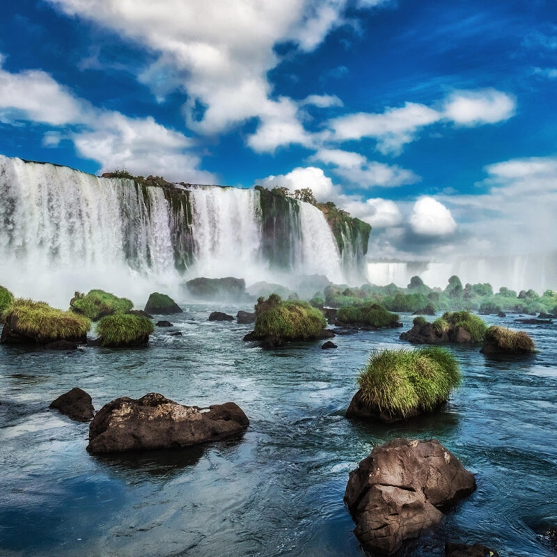 A powerful waterfall cascading into a blue river, surrounded by water-soaked black rocks and green tufts. Luxury Argentina vacations.